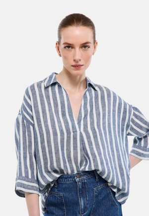 Jeune femme aux cheveux lissés en arrière portant une chemise ample à rayures verticales bleues et blanches et un jean bleu taille haute, main sur la hanche.