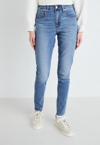 Jeans de tiro alto en azul claro con un ajuste slim, hechos de denim. Presentan bolsillos frontales y un cierre de cremallera. Combinados con zapatillas neutras.