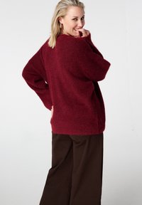 Pull oversize bordeaux en maille texturée, manches longues et coupe décontractée. Associé à un pantalon large marron.