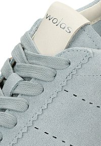 Gros plan sur une basket en daim gris clair avec des lacets assortis et une languette crème portant la marque "wojas" en noir.