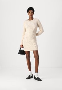 Lys beige strikket kjole med diamant kabelmønster; lange ermer, ribbestrikket hals og kant. Slim fit, brukt med svart veske og sko.