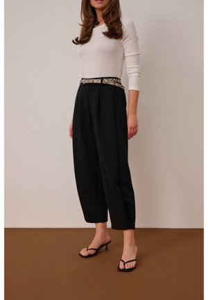 Nő, fehér, testhezálló hosszú ujjú felsőt visel, fekete magas derekú bőszárú nadrágot kígyómintás övvel, fekete magassarkú szandálban, barna padlón.