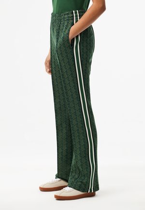 Persoon draagt groene patterned trainingsbroek met witte zijstrepen en witte sneakers met rubberen zolen, staand met één hand in de zak.