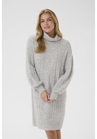 Svetlo siva, pletena obleka s turtleneck ovratnikom, ki prikazuje pletene vzorce in rebraste manšete. Tkanina se zdi mehka in teksturirana.
