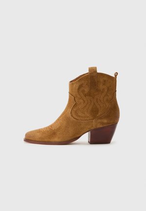 Stivale da cowboy alla caviglia in suede marrone con cuciture decorative, linguette per l'abbottonatura e tacco in legno impilato, mostrato in profilo su uno sfondo bianco.