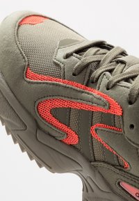 Sneakers w kolorze oliwkowym wykonane z siateczki i zamszu, podkreślone czerwonymi akcentami, z teksturowaną podeszwą i grubą, solidną konstrukcją.