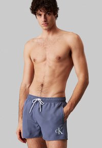 Shorts da nuoto grigi con vita elastica, stringa e tasche laterali. Presentano un logo bianco a contrasto sull'orlo.