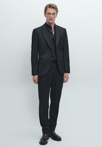 Tmavý oblek na míru vyrobený z vlněné tkaniny, obsahující strukturovanou blejzr s dvěma knoflíky, doplněný o slim-fit kalhoty a kožené boty.