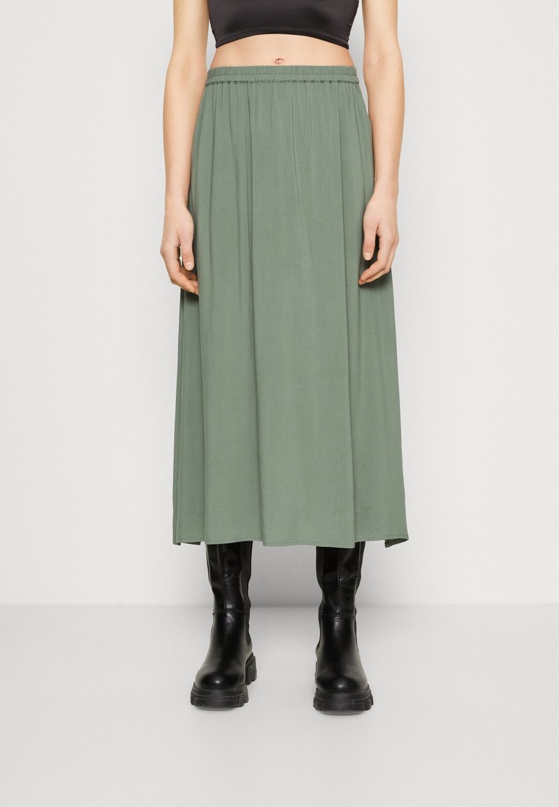 Personne portant une jupe midi taille haute vert olive avec des bottes noires épaisses, debout devant un fond clair uni.