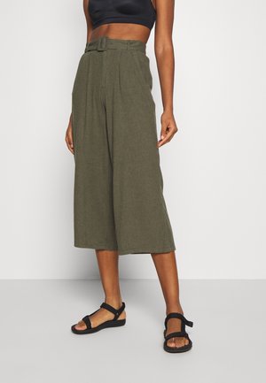 Vrouw die wijde, olijfgroene cropped broek met hoge taille en bijpassende stoffen riem draagt, gecombineerd met zwarte sandalen en een zwart cropped topje.