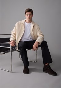 Chaqueta casual beige sobre camiseta blanca, pantalones negros y zapatos de ante marrón oscuro. Modelo sentado en una silla metálica minimalista, fondo gris.