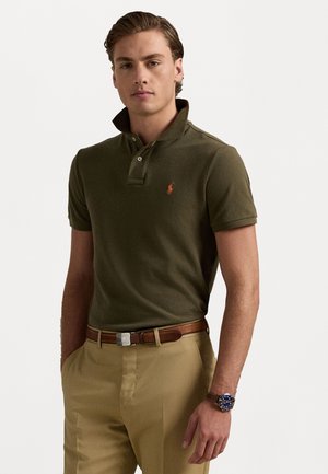 Donkergroene polo met een subtiele textuur, kraag met drie knopen en een oranje logo. Gecombineerd met lichtbeige broeken en een bruin leren riem.