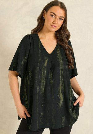 Kobieta o długich brązowych włosach, ubrana w luźną czarną bluzkę z dekoltem w serek i zielonymi pionowymi paskami, z rękami częściowo w kieszeniach, na neutralnym tle.