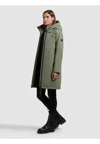 Parka verde oliva con chiusura a zip, cappuccio e maniche lunghe. Presenta una toppa sul braccio superiore e tasche laterali con zip. Abbinata a stivali neri.