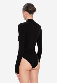 Kobieta z włosami w kok, ubrana w czarny bod bodysuit z długim rękawem, odwrócona plecami z rękami luźno opadającymi wzdłuż ciała na jednolitym tle.