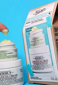 Produkty do pielęgnacji skóry Kiehl's w białych słoikach; kremowa kuracja pod oczy z zielonym tekstem, poniżej ultra nawilżający krem. Tło z tektury kartonowej z niebieskimi akcentami.
