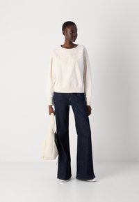Felpa crema con motivo floreale testurizzato sul giromanica, abbinata a jeans a gamba larga blu scuro. Il modello tiene una borsa color beige chiaro.