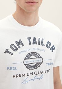 Weißes Baumwoll-T-Shirt mit dunkelblauem "TOM TAILOR"-Logo in einem runden Emblem, das "seit 1962" und den Text "PREMIUM QUALITÄT" in Schwarz zeigt.