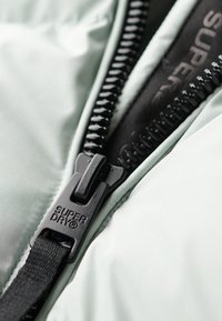 Chaqueta de color verde claro con cremallera y tejido. La cremallera tiene un tirador texturizado en negro que muestra la marca "SUPER DRY". Material suave con un diseño elegante.