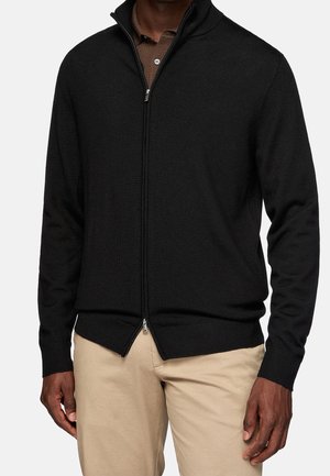 Homme portant un pull zippé noir par-dessus une chemise marron à col et un pantalon beige, debout devant un fond clair uni.