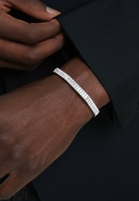 Silberarmband mit einer schlanken, länglichen Form, das strukturierte Rillen und eine glatte Oberfläche aufweist, getragen an einem dunkel gekleideten Handgelenk.