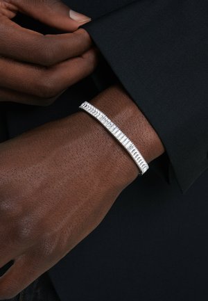 Zilveren armband met een slanke, langwerpige vorm, gekenmerkt door gestructureerde ribbels en een gladde afwerking, gedragen om een donker geklede pols.