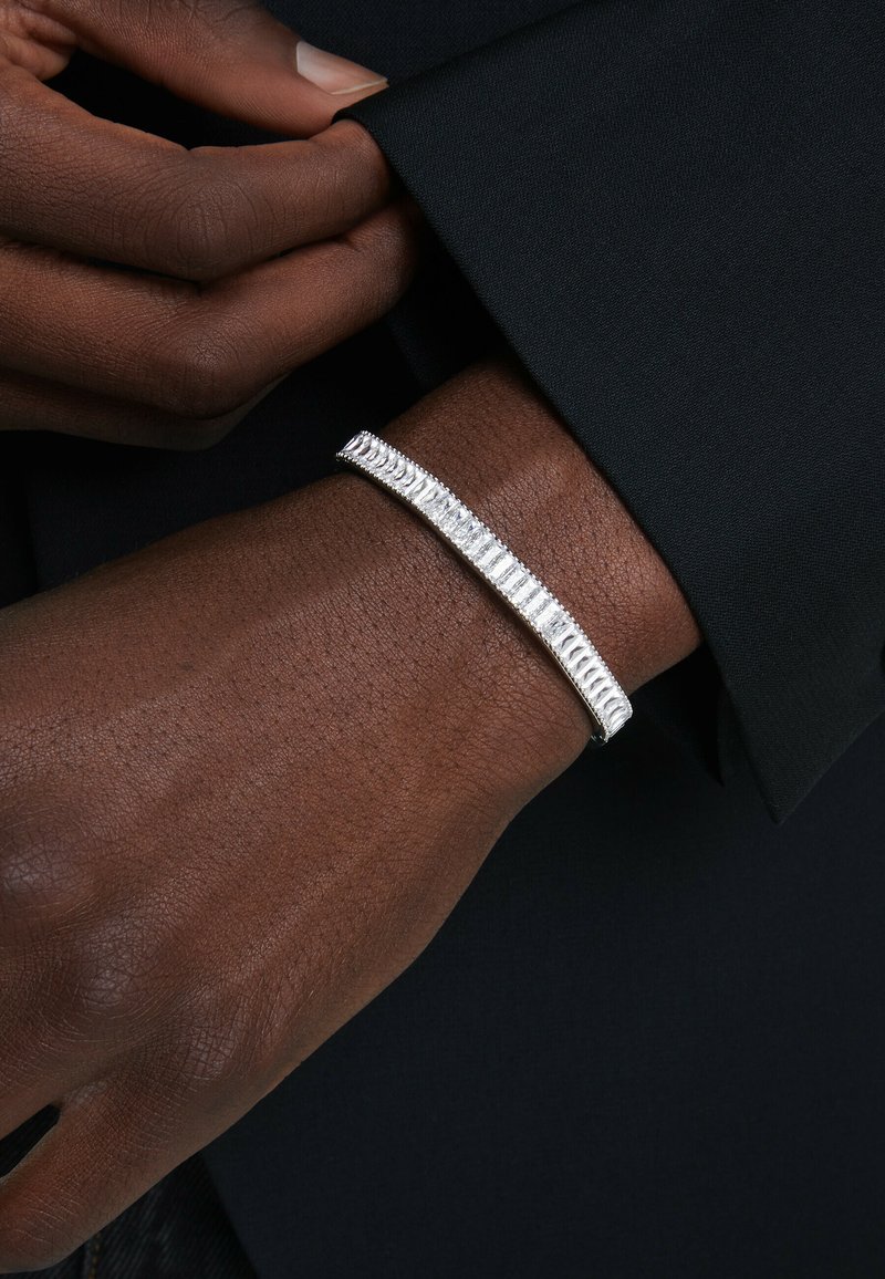 Silberarmband mit einer schlanken, länglichen Form, das strukturierte Rillen und eine glatte Oberfläche aufweist, getragen an einem dunkel gekleideten Handgelenk.