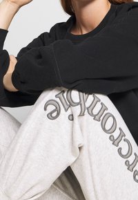 Sudadera negra con puños acanalados y un ajuste relajado, combinada con pantalones de chándal gris claro que presentan un texto impreso oscuro a lo largo de la pierna.
