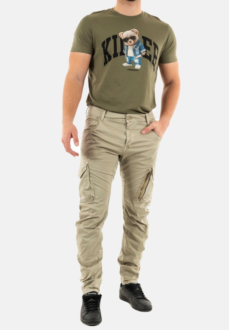 T-shirt vert olive avec un graphique d'ours, associé à un pantalon cargo beige clair avec des poches zippées et une coupe ample, complété par des baskets noires.