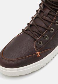 Bruine leren sneaker met een gestructureerd oppervlak, oranje stiksels, metalen oogjes en bruine veters. Witte rubberen buitenzool.