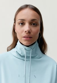 Sudadera de cuello alto azul claro con cordones en la parte delantera y detalle del logo en el pecho. Textura de tela suave, diseño ajustado, estilo minimalista.