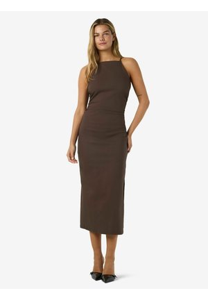 Femme portant une robe midi sans manches, cintrée, de couleur brun foncé avec un col carré et des talons noirs à bout pointu, debout devant un fond uni.