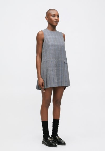 Pelēkā svītraina kleita bez piedurknēm, ar plisētu svārku un sānu kabatu. Melni loaferi ar zeltītu aparatūru un melnas zeķes.