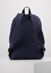 Sac à dos en tissu bleu marine avec des bretelles rembourrées, une poignée supérieure et une texture lisse. Présente un design minimaliste et sans motifs visibles.