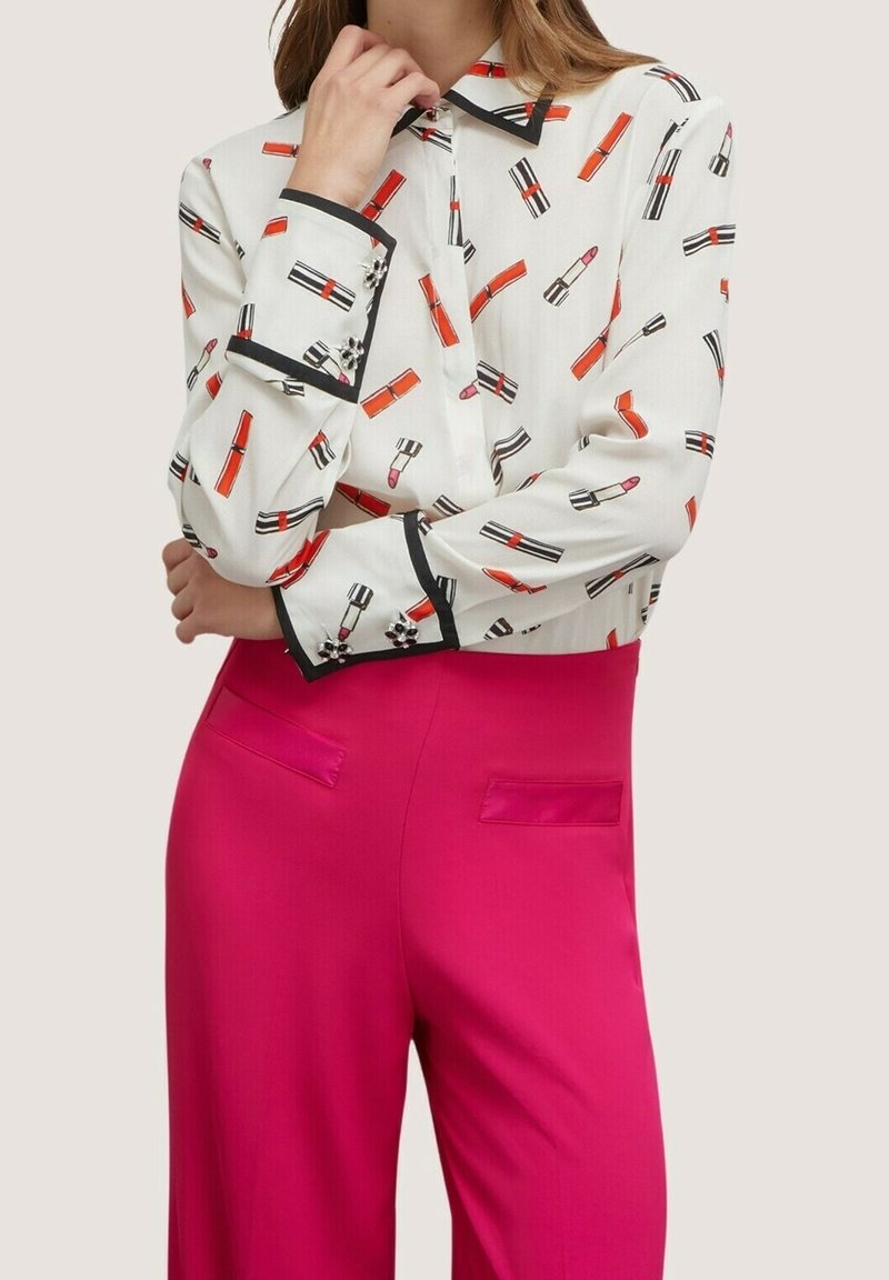 Blouse blanche avec un motif répétitif de rouge à lèvres en rouge et noir, dotée d'un col noir. Associée à un pantalon rose vif, les deux étant cintrés.