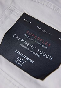 Tapered fit clothing tag on a light-colored fabric, featuring black text detailing "Superflex," "Cashmere Touch," and "Lindbergh 1927."