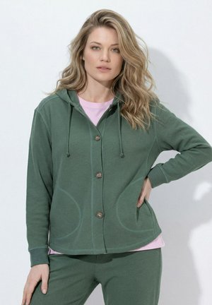 Frau mit blondem, welligem Haar, die einen grünen, geknöpften Hoodie und passende Hose trägt, eine Hand in der Hüfte und darunter ein hellrosa Shirt.