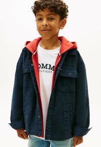 Granatowa kurtka z korduroju z dwoma przednimi kieszeniami, nałożona na czerwoną bluzę z kapturem i białą koszulkę z czarnym napisem. Luźne dżinsy.