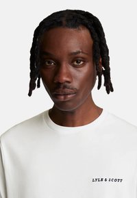 Camiseta blanca de manga corta con cuello redondo y un logotipo bordado en negro en el lado izquierdo del pecho. Tejido suave con textura mínima.