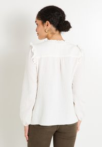 Blusa blanca de manga larga hecha de una tela texturada, con acentos de volantes en los hombros y un sutil diseño de pliegues en la parte trasera.
