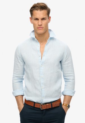 Camisa abotonada de rayas azul claro con cuello esparcido, mangas largas arremangadas en los puños, combinada con un cinturón de cuero marrón y pantalones azul marino.