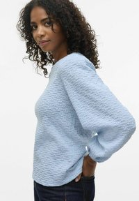 Top a maniche lunghe di un blu chiaro testurizzato con una vestibilità rilassata e scollatura rotonda, caratterizzato da motivi sottili. Indossato con jeans scuri.