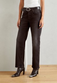 Jeans di denim nero con taglio dritto, cinque tasche e cintura con fibbia argentata. Stivali alti marroni con punte metalliche.