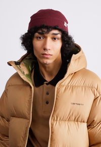 Tan puffer jacket with a green interior, paired with a brown polo shirt. Model wears a maroon beanie and has a septum piercing.