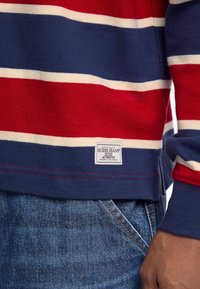 Prugasta košulja dugih rukava u crvenoj, plavoj i bijeloj boji od pamuka s malom oznakom "Guess Jeans" na donjem rubu, u kombinaciji s trapericama.