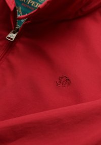 Rode zip-up jas met geborduurd "JD" logo op de borst en gedeeltelijk zichtbare groene binnenlabel met gouden tekst.