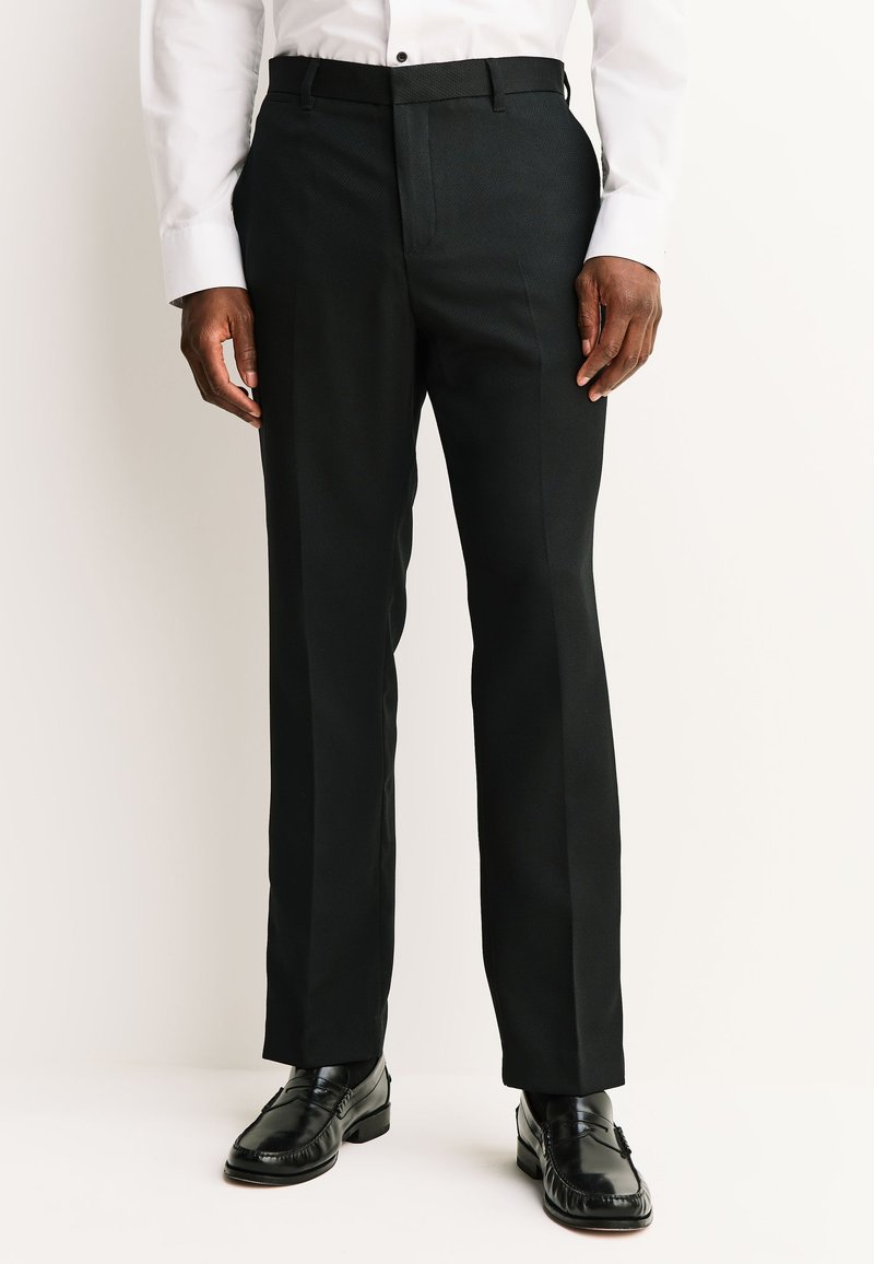 Homme portant un pantalon de costume noir ajusté, des mocassins en cuir noirs et une chemise blanche à boutons sur un fond uni clair.