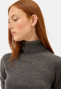 Grauer Wollrollkragenpullover mit ribbeliger Kragenkante, der eine weiche Textur und ein tailliertes Design aufweist, vor einem neutralen Hintergrund präsentiert.
