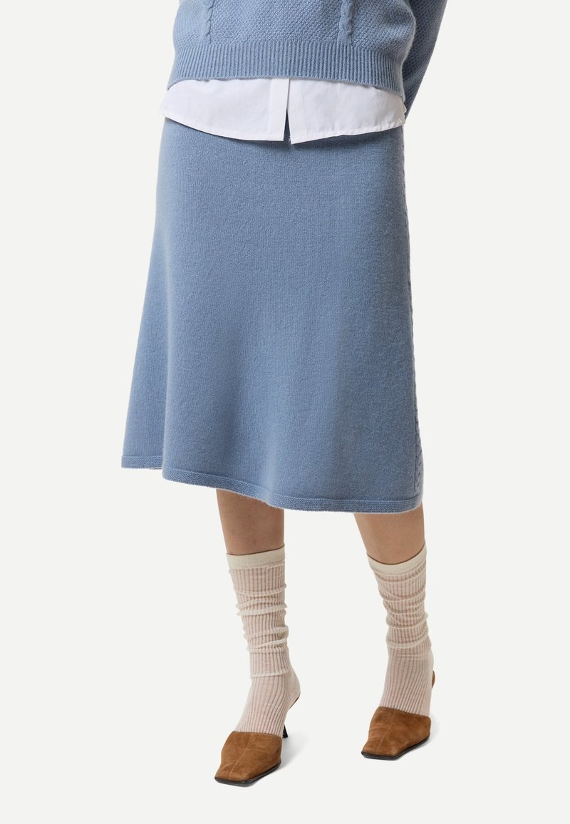 Kvinna klädd i en ljusblå stickad midikjol, vit skjorta, matchande blå tröja, krämfärgade ribbade strumpor och bruna klackskor.