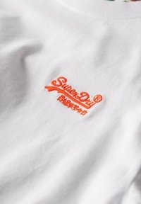 Tela blanca con pequeño logo naranja de "SuperDry" y caracteres japoneses bordados en la superficie.